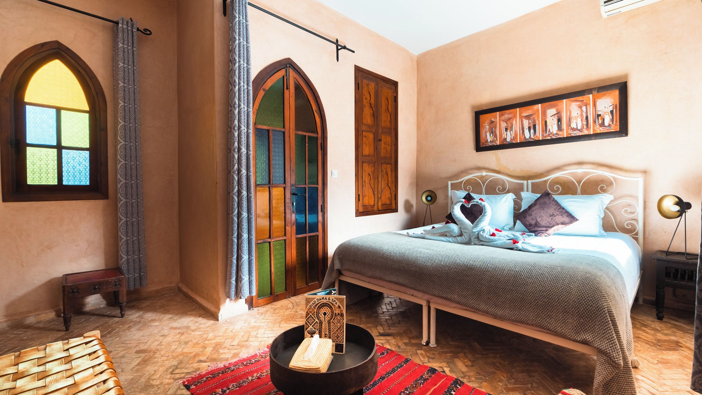 Ein Schlafzimmer mit einem Bett, einem kleinen Tisch, einem Sessel, einem Wandbild und einem Fenster mit gemusterten Vorhängen.
