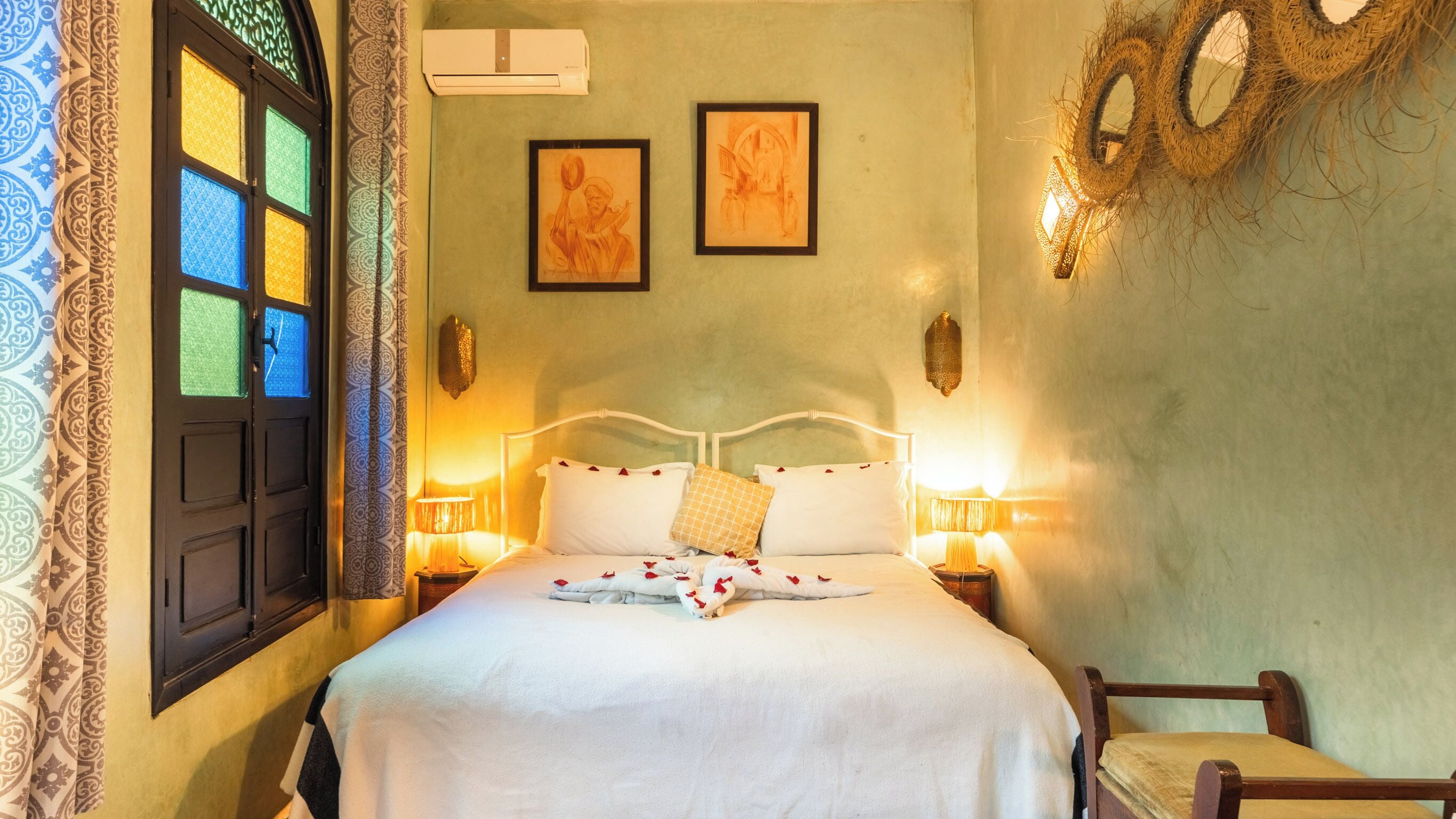 Une chambre à coucher comprenant un lit, deux tables de chevet, une chaise, une fenêtre avec des rideaux à motifs et deux œuvres d’art encadrées accrochées au mur.