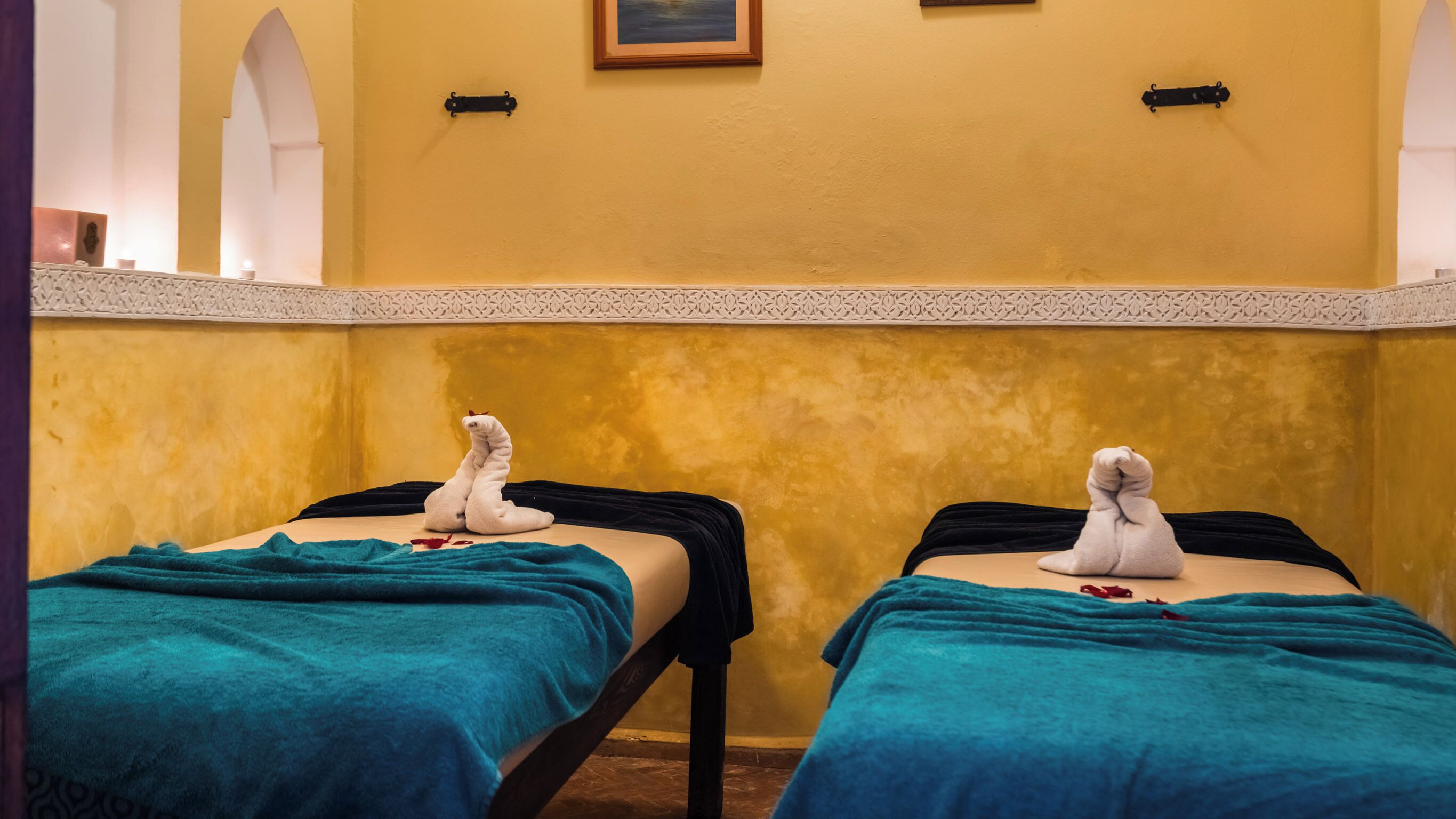 Dos camillas de masoterapia con toallas y pétalos de rosa, en una habitación con paredes amarillas y un detalle decorativo.