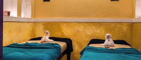 Dos camillas de masoterapia con toallas y pétalos de rosa, en una habitación con paredes amarillas y un detalle decorativo.