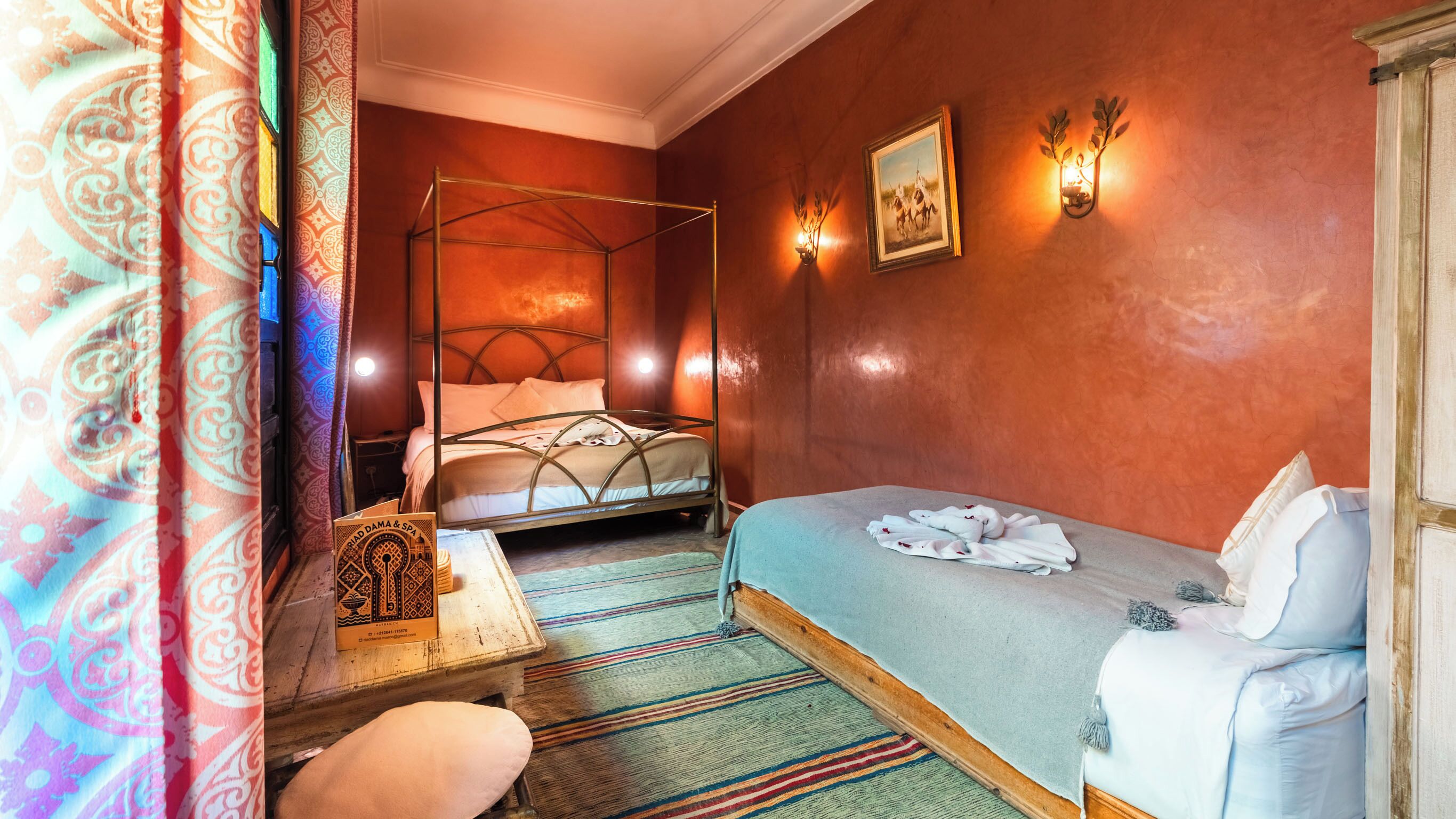 A room with a four-poster bed, a bedside table with a book, a wall-mounted light, and a framed picture.