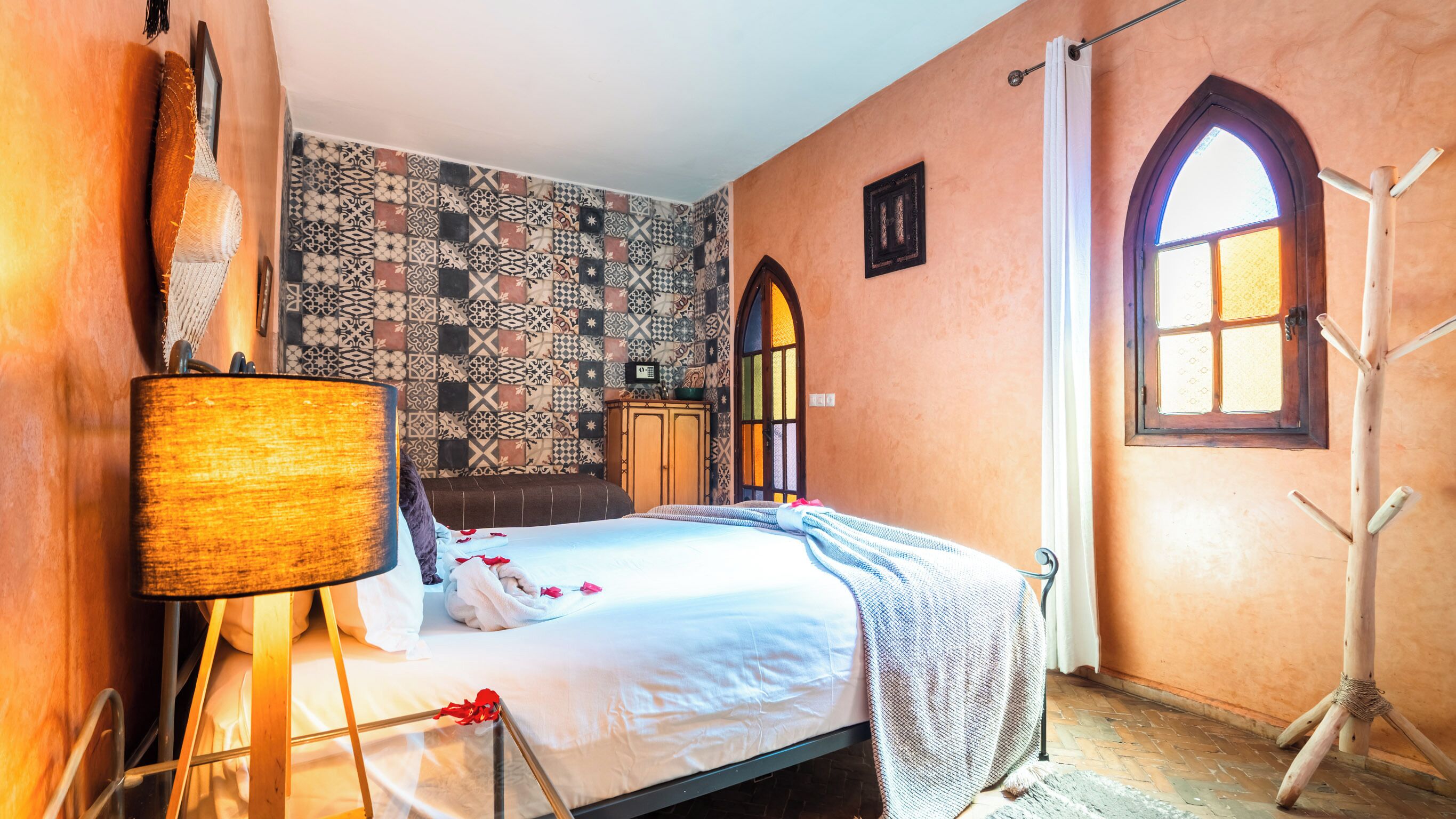 A bedroom with a patterned wall, a bed with white linens, a wooden wardrobe, and a window with a view.