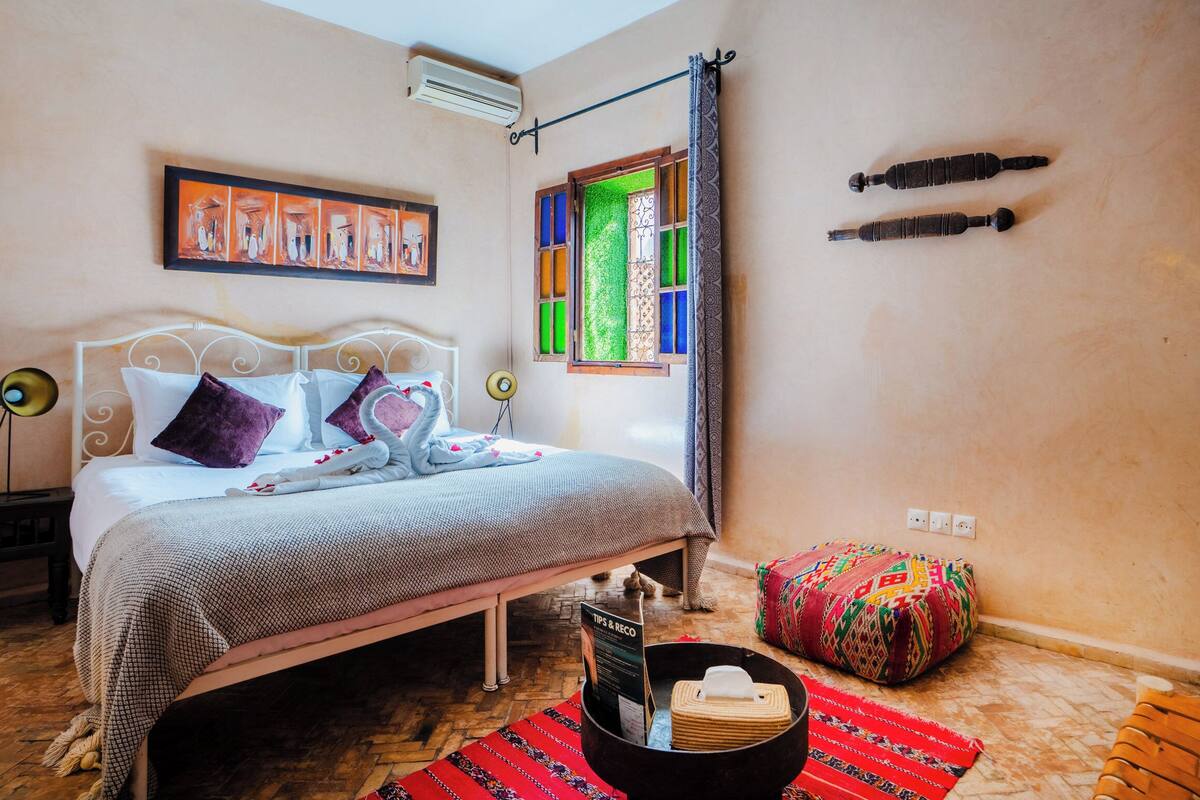 A bedroom with a bed, a window with colorful panes, a radiator, and a wall-mounted air conditioner.