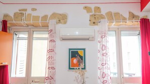 A room with a stone wall, air conditioning unit, windows with red curtains, and a framed picture on the wall. - Yenne Home (Cagliari)
