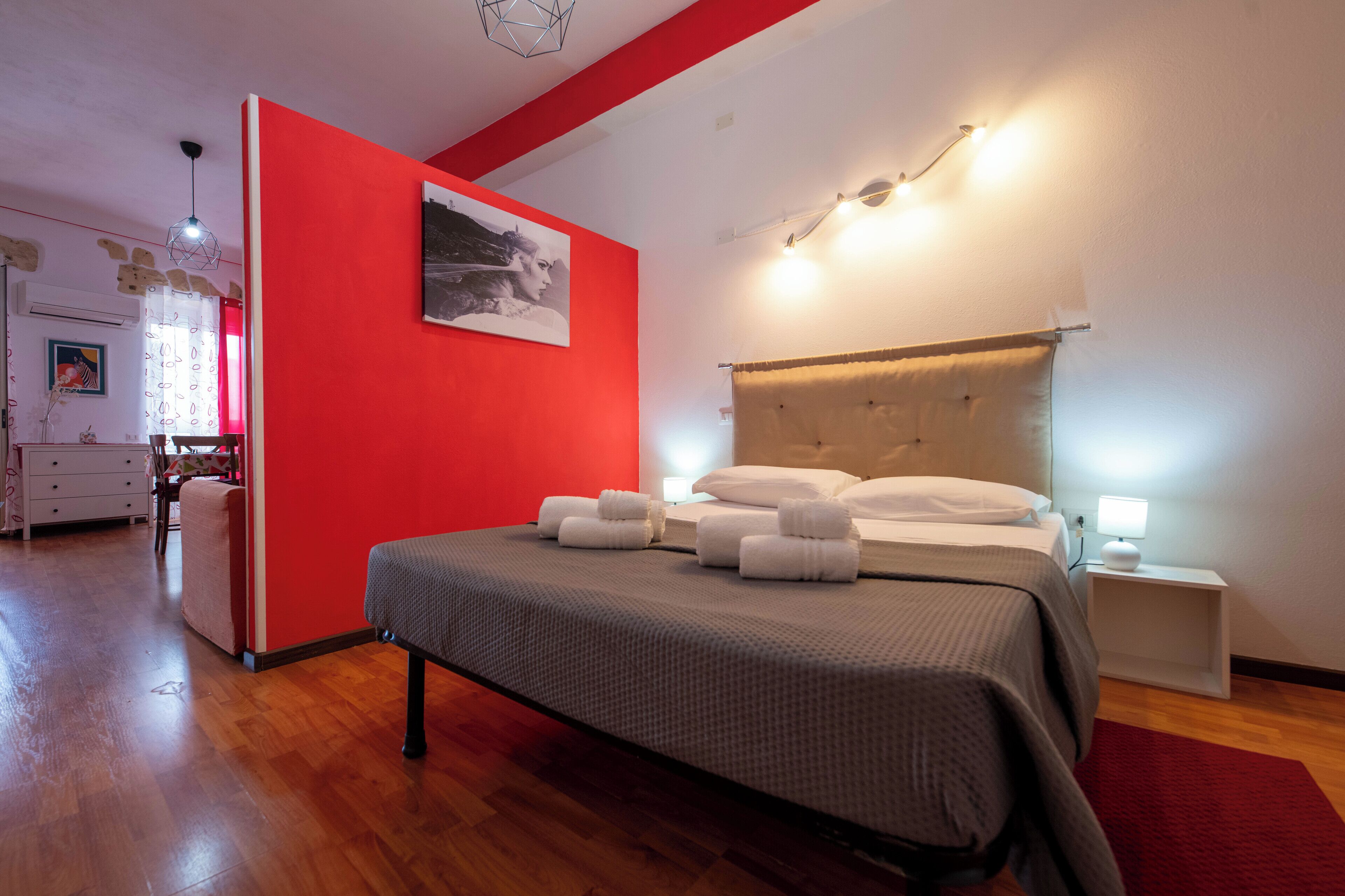 A hotel room with a red accent wall, a large bed with neatly folded towels, a bedside table with a lamp, and a modern ceiling light fixture.