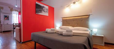 A hotel room with a red accent wall, a large bed with neatly folded towels, a bedside table with a lamp, and a modern ceiling light fixture.