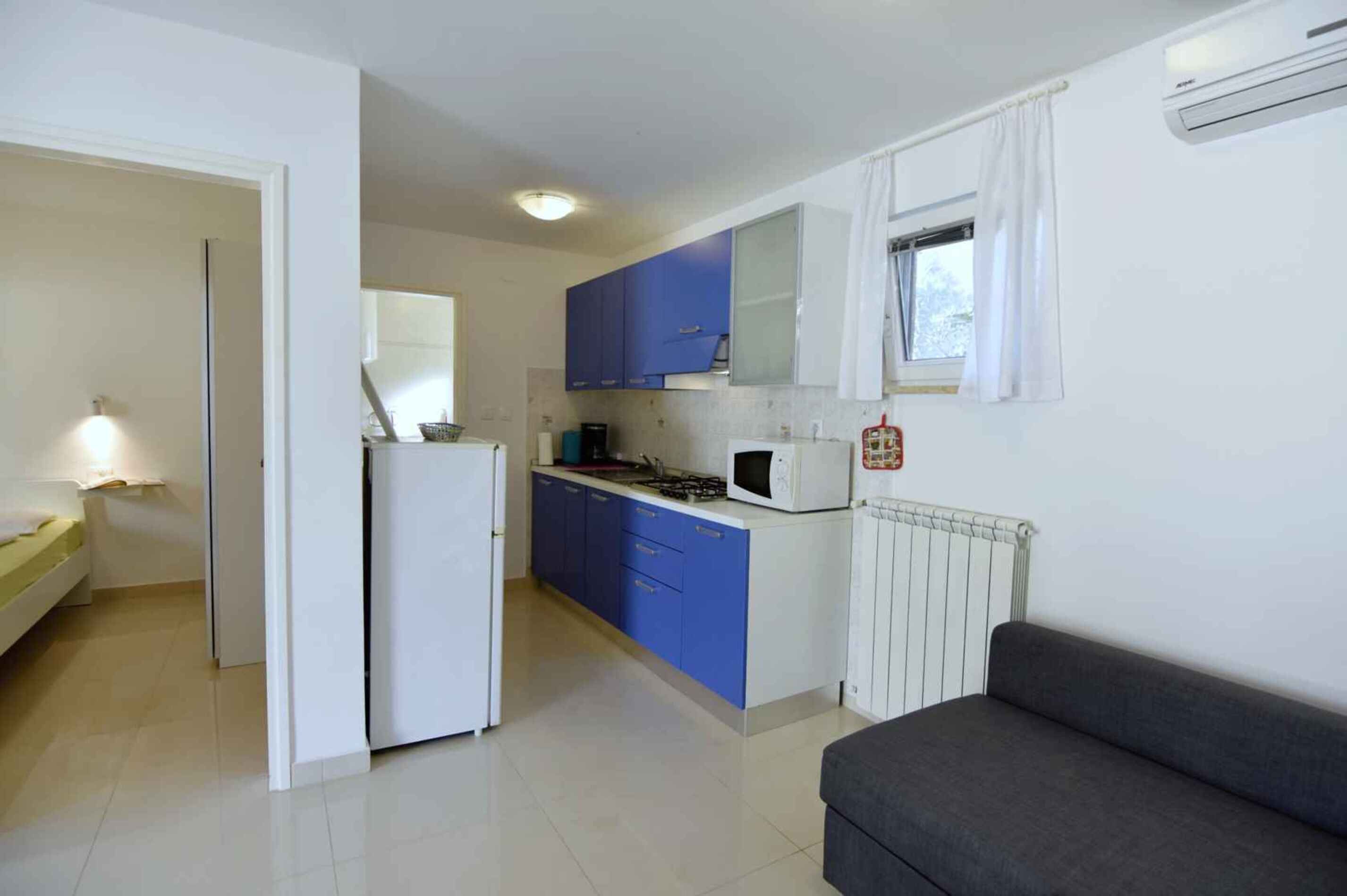 Cuina compacta amb armaris blaus, un refrigerador blanc, microones i una finestra amb cortines.
