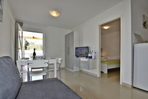 Ein modernes Wohnzimmer mit grauem Sofa, einem Esstisch mit Stühlen, einem Fernseher und einer kleinen Küchenzeile.