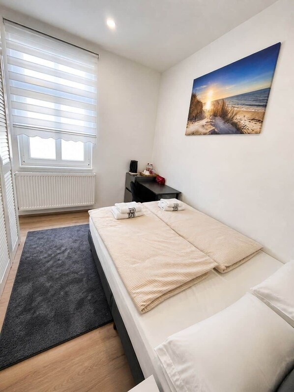 A bedroom with a bed, a desk, a chair, a radiator, a window with blinds, and a painting on the wall.