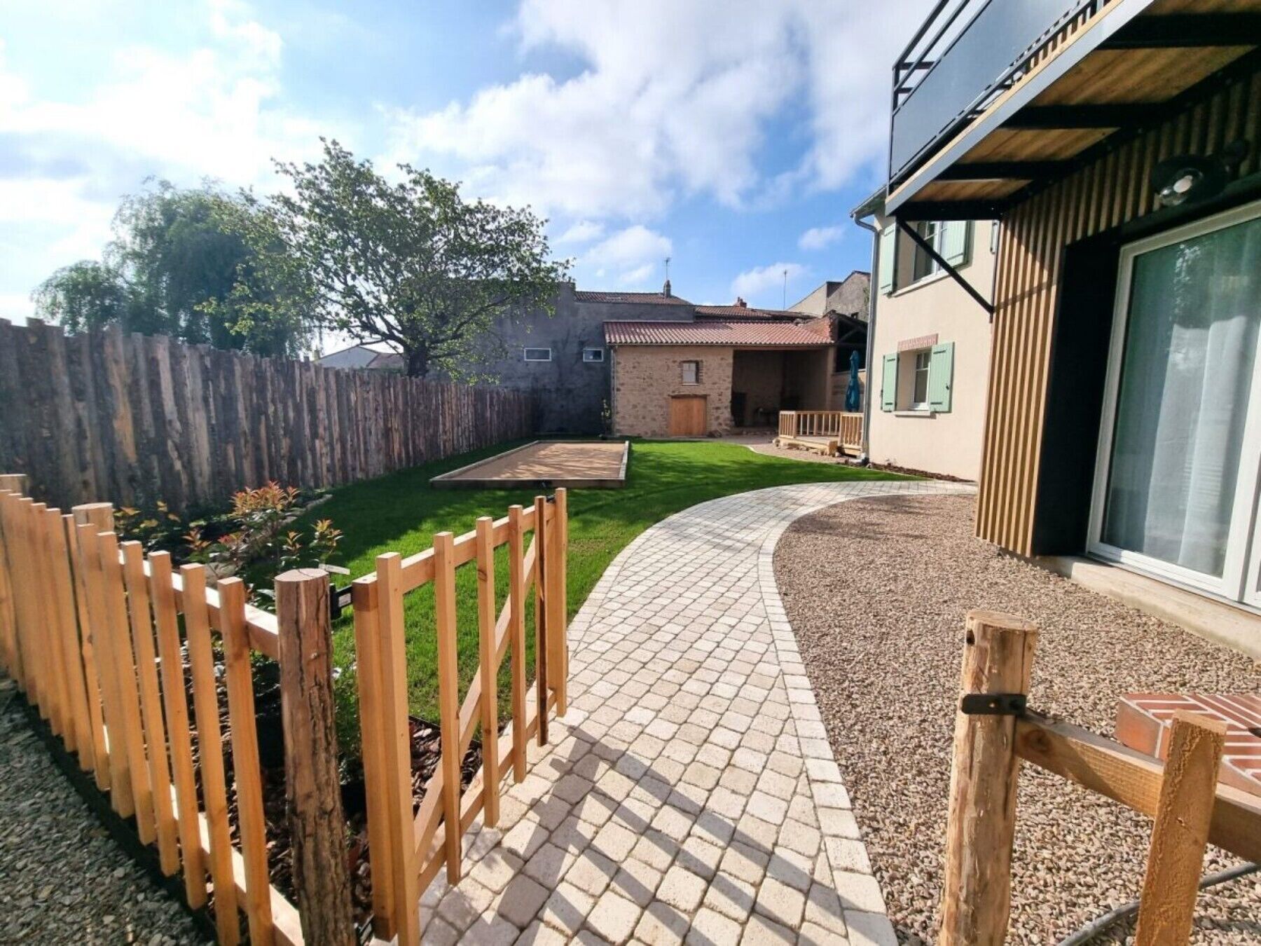 A residential property with a paved driveway, a wooden fence, a grassy lawn, and a brick patio area.