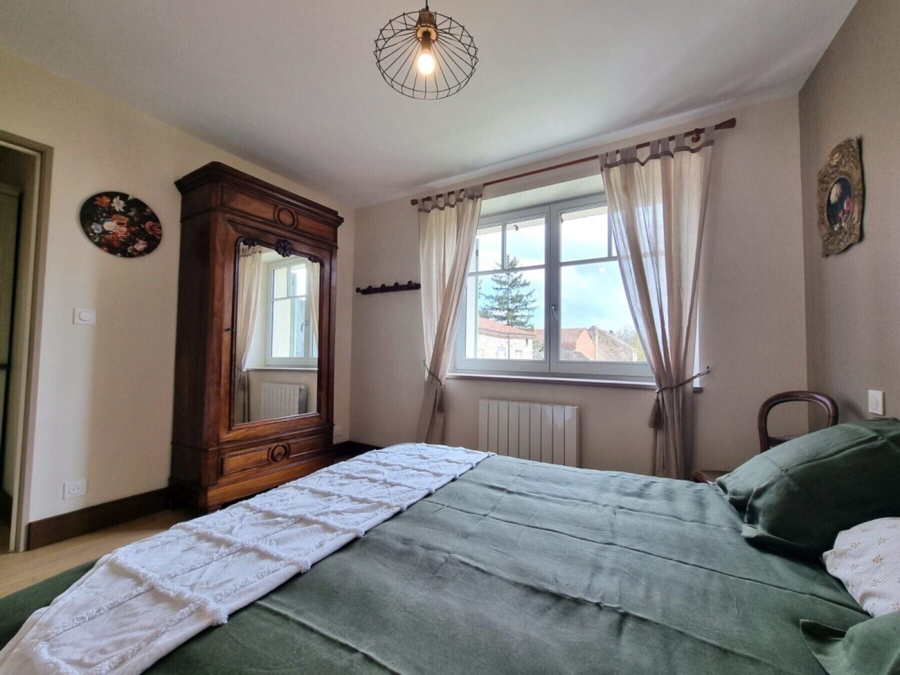 A bedroom with a bed, a dresser, a window with curtains, a radiator, and a wall clock.
