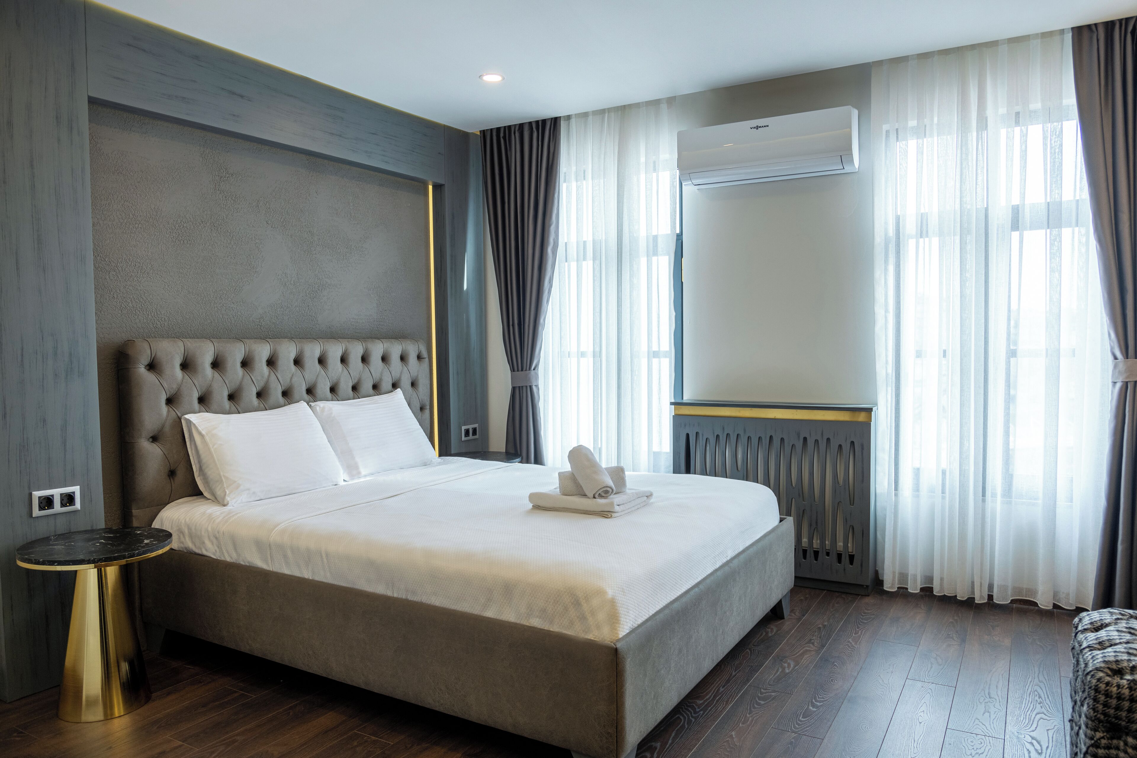 A neatly made bed with white linens and a folded towel, a round gold side table, and a large window with sheer curtains.