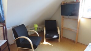 Una habitación con dos sillas negras, una mesita con lámpara y un televisor sobre un soporte de madera.