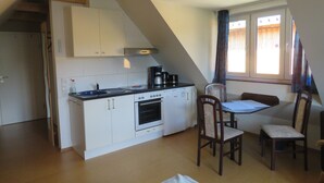 Cocina compacta con armarios blancos, estufa y cafetera. Hay una mesa de comedor con dos sillas y una ventana con cortinas.