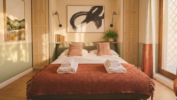 A well-lit bedroom with a large bed, a painting, a potted plant, and a window with curtains. - CS Trastevere Glorioso Signature Apt. (Rome)