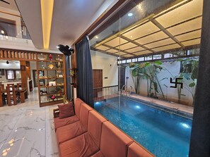 Un espace piscine intérieur moderne doté d’un mur en verre, d’un coin salon avec un canapé et d’un espace repas avec une table et des chaises en bois.