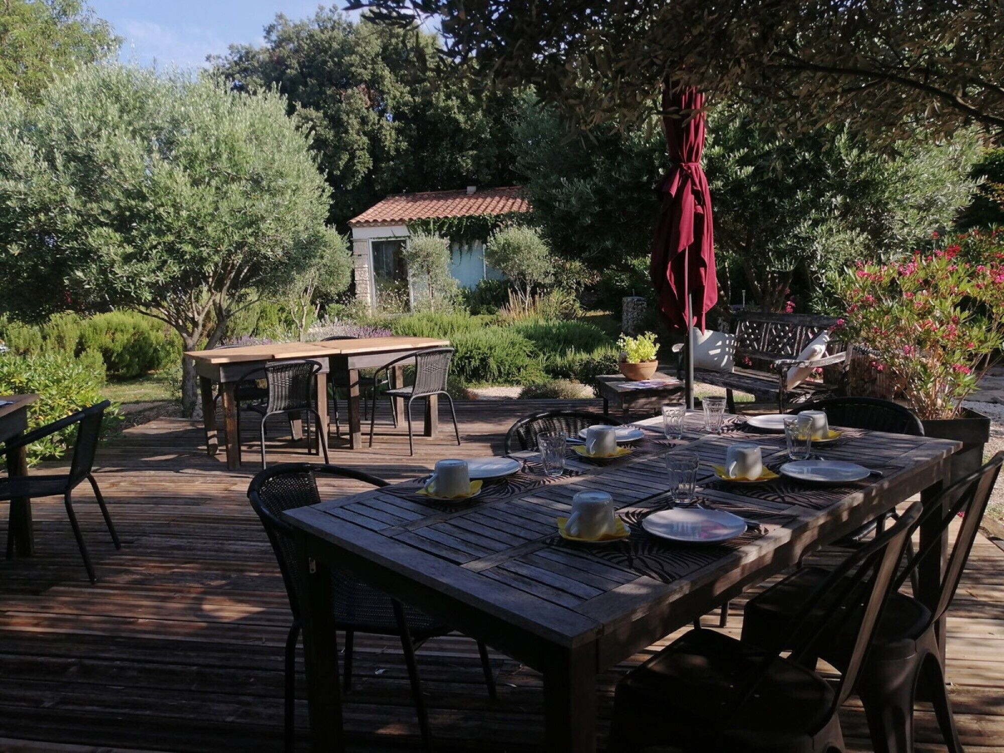 An outdoor dining area with wooden tables and chairs, surrounded by lush greenery and a small building in the background.
