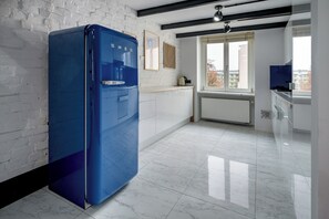 Une cuisine moderne avec un réfrigérateur bleu, des armoires blanches et une fenêtre donnant sur des immeubles.