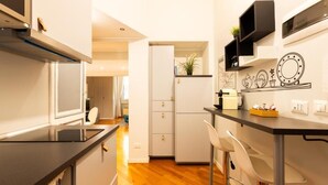 A compact kitchen with a black countertop, white cabinets, and a refrigerator. There are two white bar stools and a small shelf with decorative items.