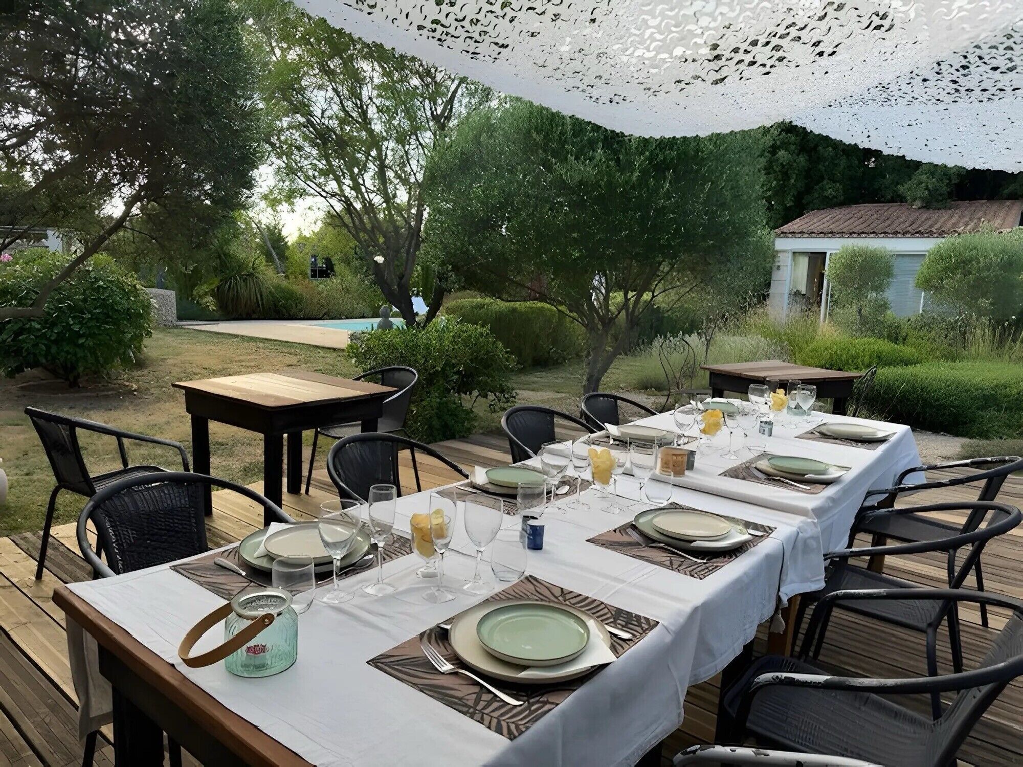 En udendørs spiseplads med borde dækket til et måltid, omgivet af vegetation og en pool.