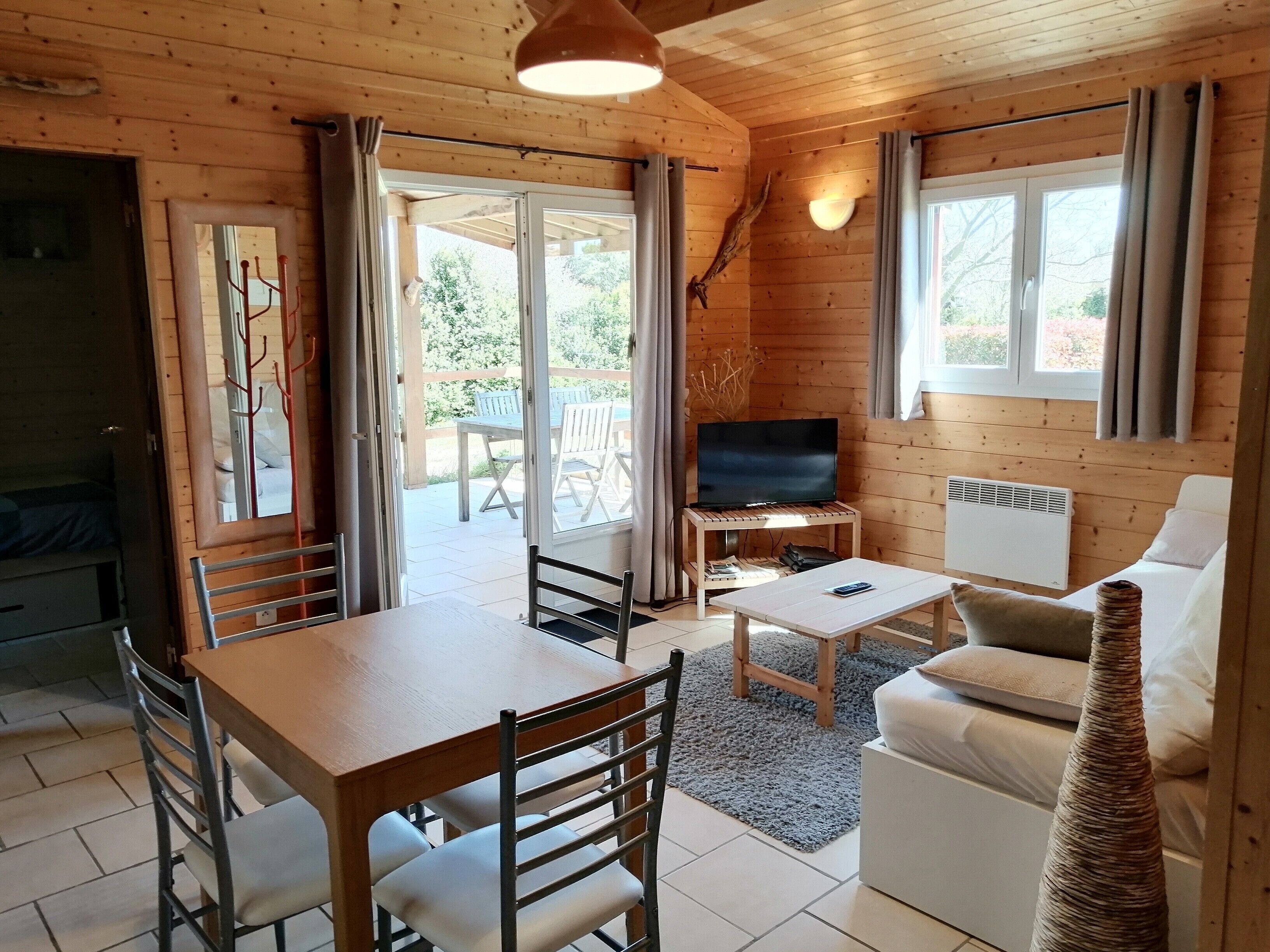 Een houten cabin met een eettafel, stoelen, een bank, een televisie op een standaard en een groot raam.