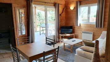 Een houten cabin met een eettafel, stoelen, een bank, een televisie op een standaard en een groot raam.