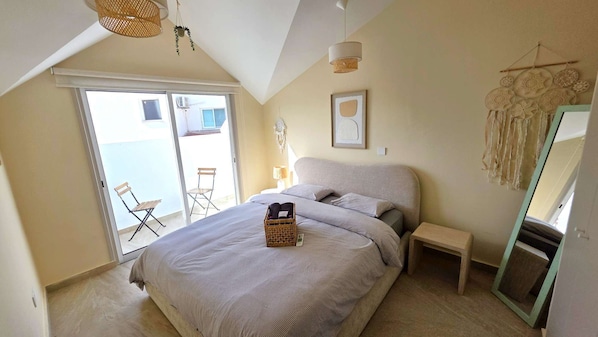 A bedroom with a large bed, a bedside table, a chair, a hanging light fixture, and a view of the outside. - Warm Design 3BR • Beach 7-min walk • Peaceful (Larnaca)