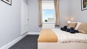 A bedroom with a bed, a window with curtains, a radiator, and a small potted plant. - Ideal Work Stays | 3BR | Free Parking | Sleeps 6 (Falkirk)