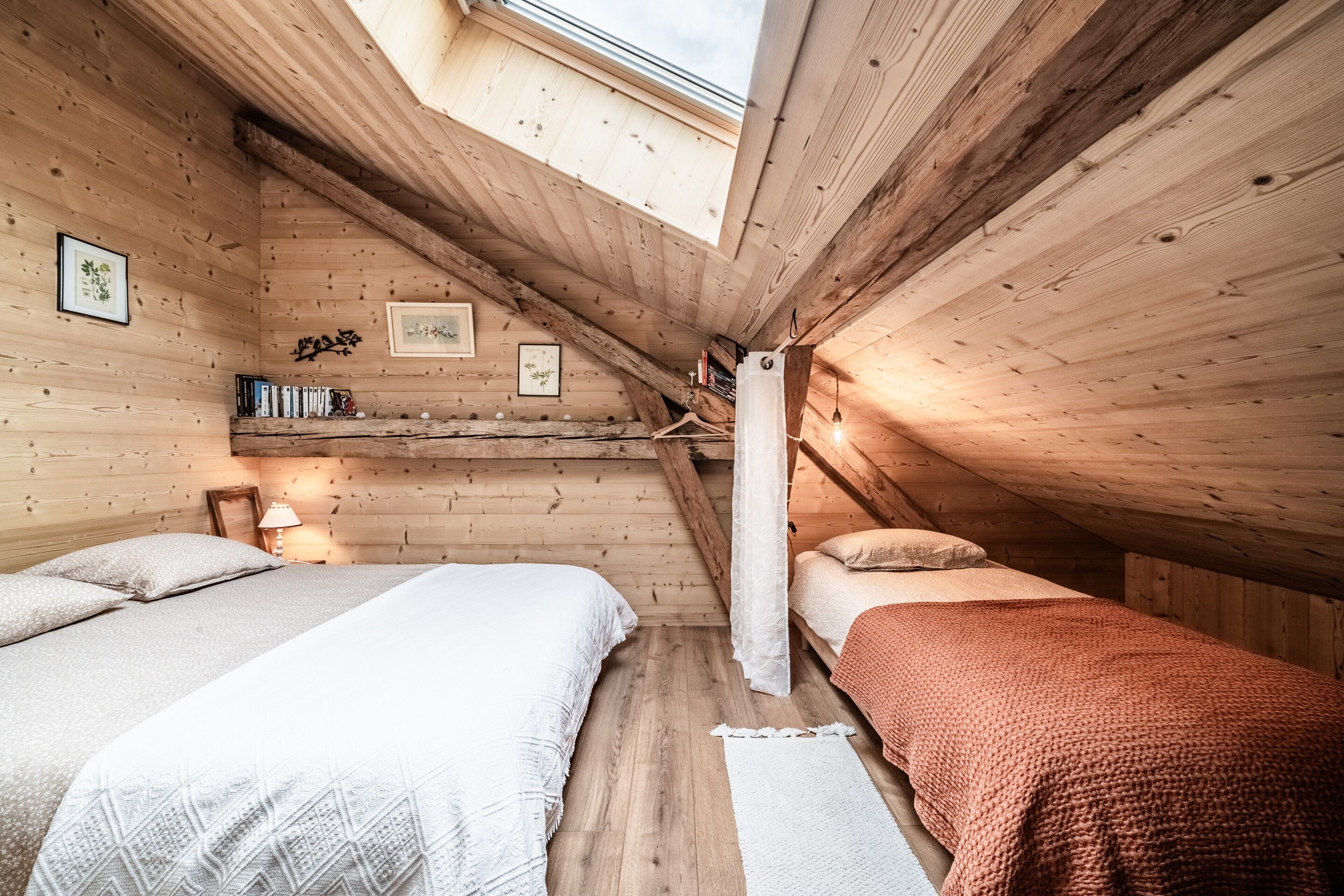 A cozy attic bedroom with two beds, wooden walls, a skylight, and a small shelf with books.