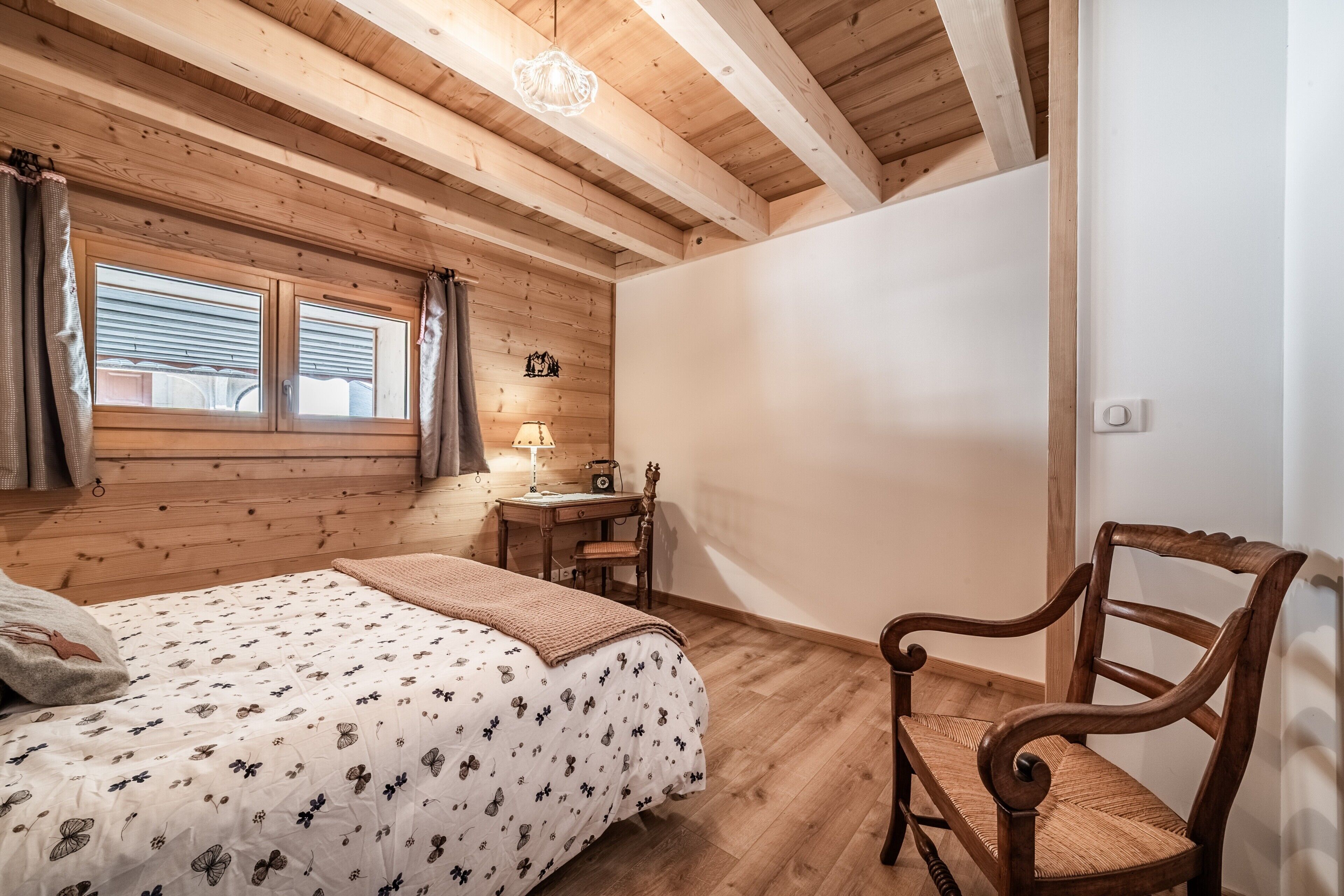 Een slaapkamer met een houten plafond, een bed met een bloemenprint, een houten stoel, een klein tafeltje met een lamp en een raam met gordijnen.