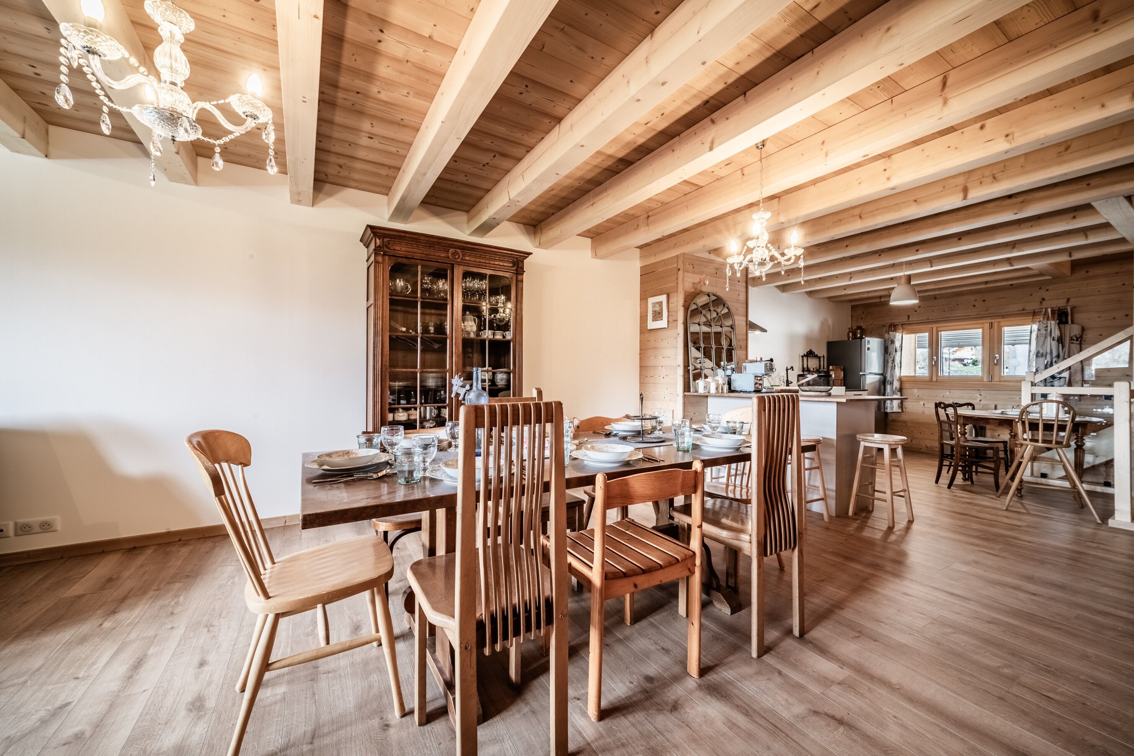 Un coin repas avec des chaises et une table en bois, un buffet en bois doté de portes en verre, et un espace cuisine avec des poutres apparentes et un lustre.