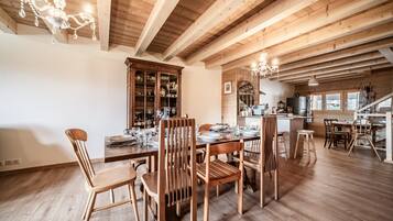 Un coin repas avec des chaises et une table en bois, un buffet en bois doté de portes en verre, et un espace cuisine avec des poutres apparentes et un lustre.
