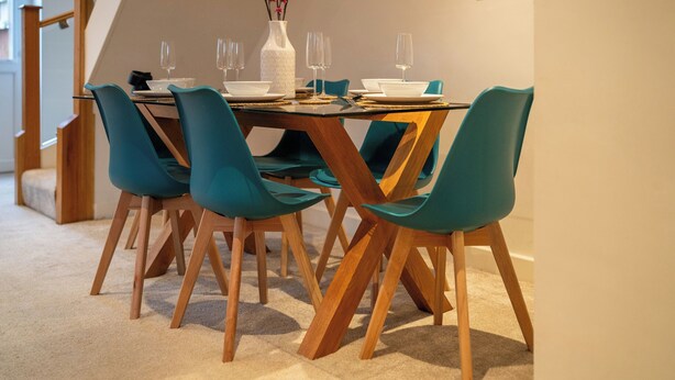A dining area with a glass table, teal chairs, and a white vase with pink flowers. - 4 Bed House - Cheltenham & Gloucester - Sleeps 8 (Gloucester)