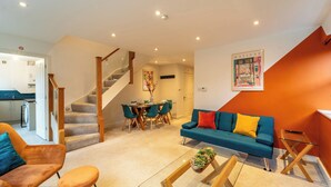 A modern living room with a blue sofa, orange armchair, and a glass coffee table. A staircase leads to a dining area with a table and chairs. - 4 Bed House - Cheltenham & Gloucester - Sleeps 8 (Gloucester)