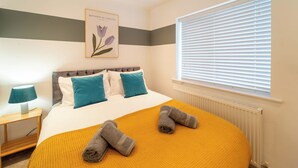 A neatly made bed with a yellow blanket, a framed botanical garden picture on the wall, and a window with blinds. - 4 Bed House - Cheltenham & Gloucester - Sleeps 8 (Gloucester)