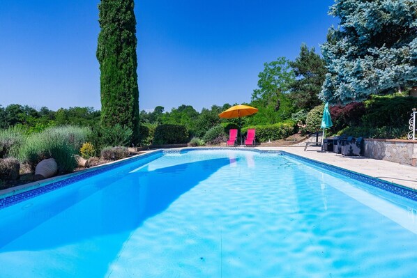 A swimming pool with a tiled edge, surrounded by greenery and a clear sky.