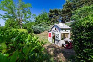 Une petite maison de jeu en bois, avec une porte rouge et un banc, entourĂ©e dâune vĂ©gĂ©tation luxuriante et dâarbres.