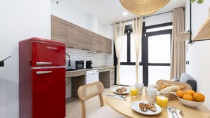 A compact kitchen with a red refrigerator, wooden cabinets, a dining table with breakfast items, and a view through a large window. - Travel Habitat Nou Mestalla 2 (Valencia)