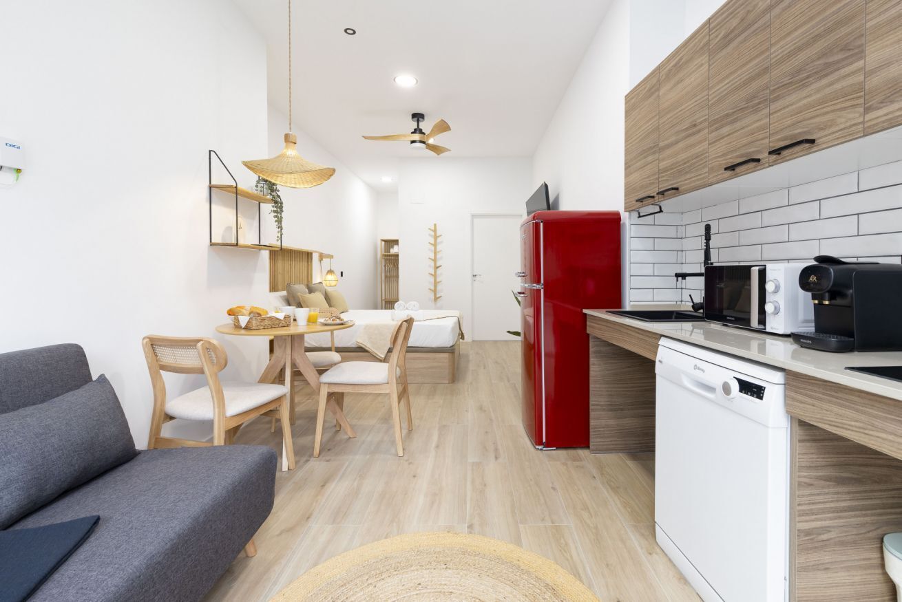 Una cocina moderna con un refrigerador rojo, lavavajillas blanco y armarios de madera. Hay una zona de comedor con una mesa y sillas, y un sofá gris en primer plano.