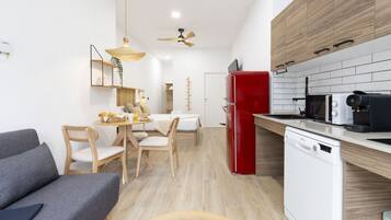 Una cocina moderna con un refrigerador rojo, lavavajillas blanco y armarios de madera. Hay una zona de comedor con una mesa y sillas, y un sofá gris en primer plano.