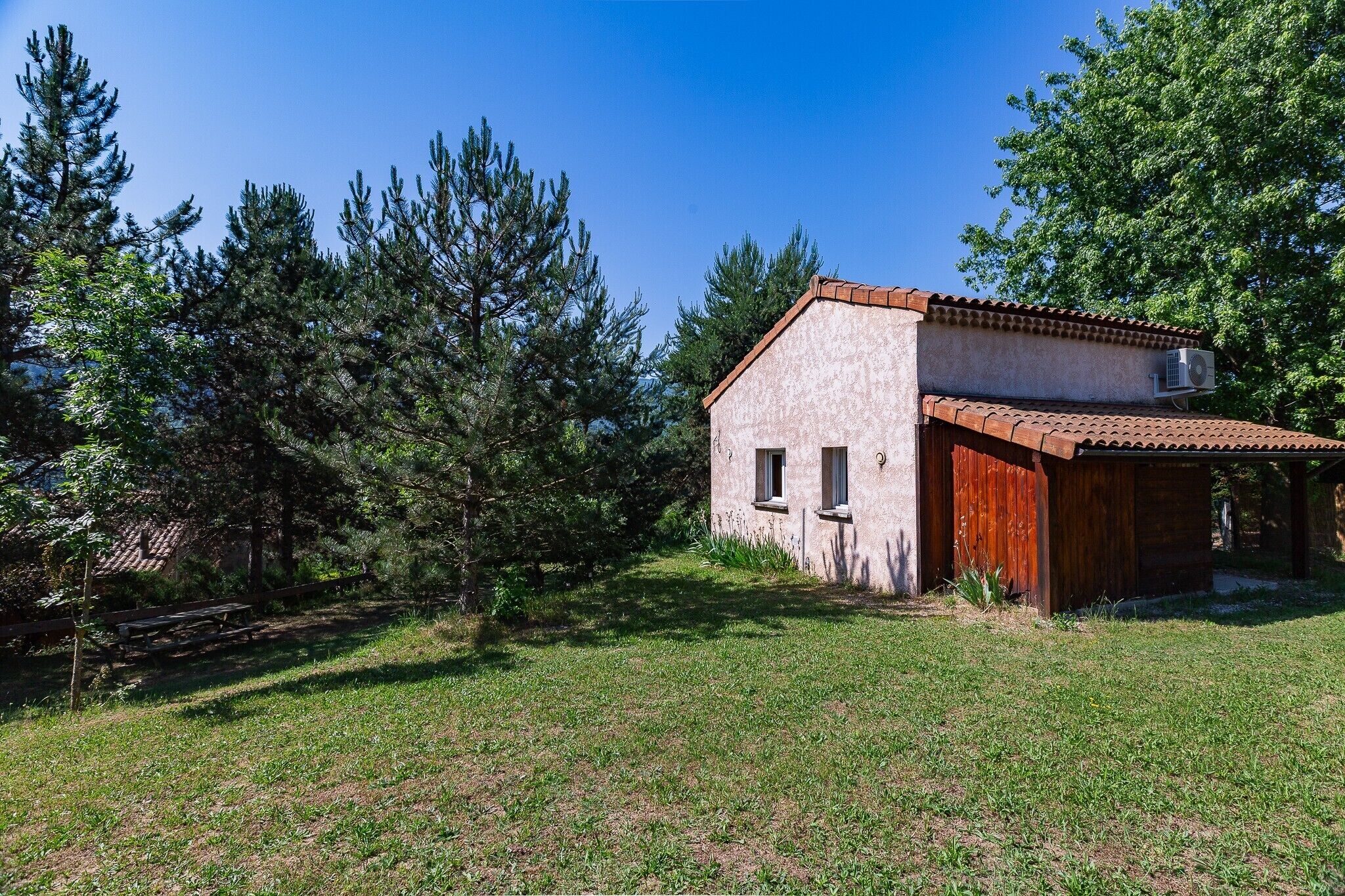 A small, single-story building with a sloped roof, surrounded by a grassy area and dense trees.