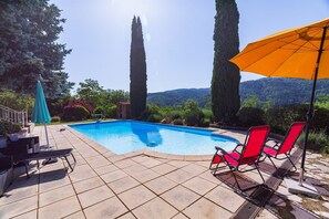 Une piscine entourée d’arbres et de collines, avec des chaises longues et un parasol.