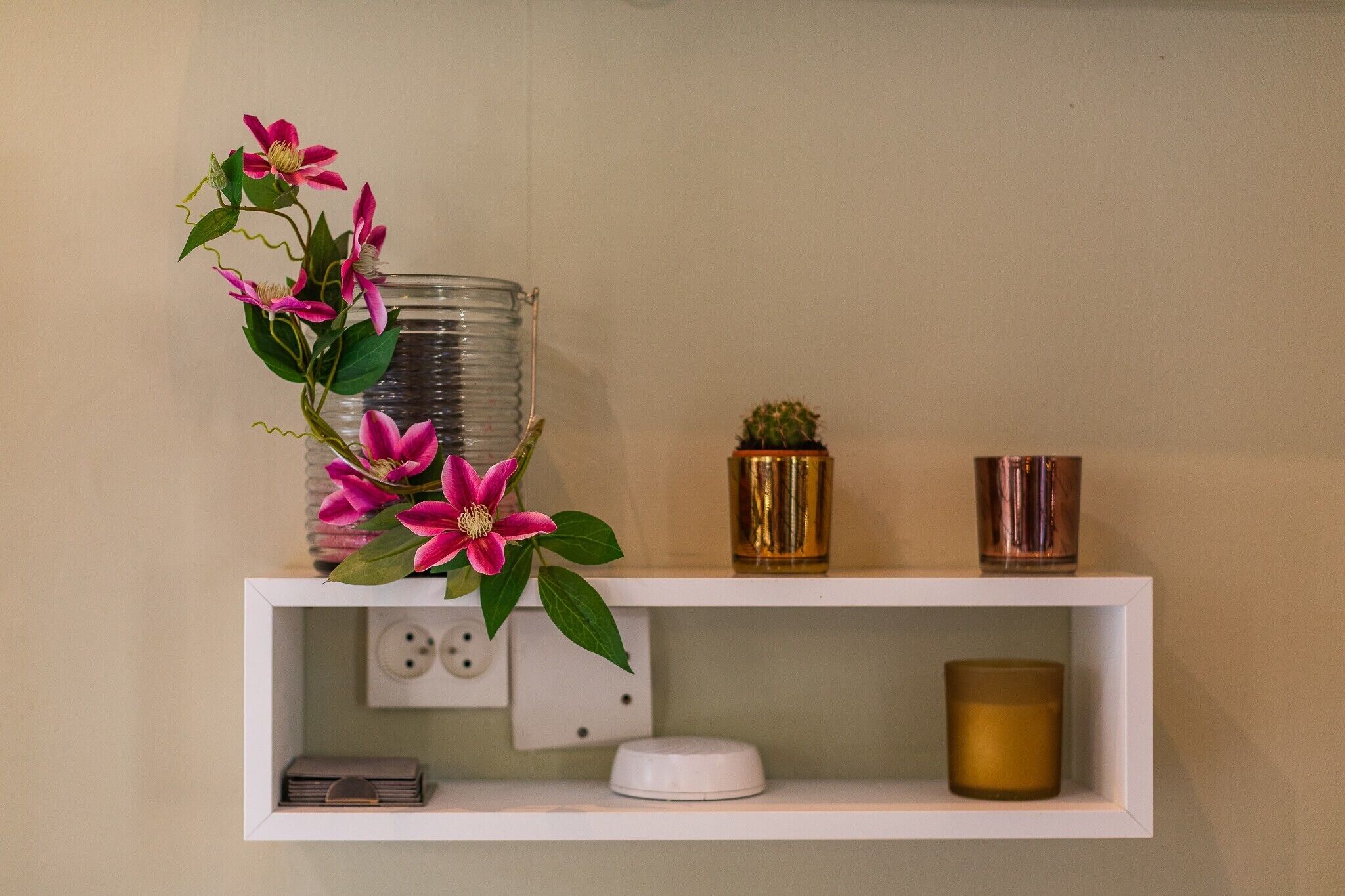 Una estantería con un florero de flores rosas, una planta pequeña en maceta y dos recipientes cilíndricos.