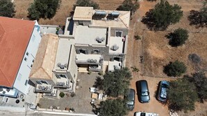 An aerial view of a residential area with multiple buildings, including a house with a red roof and a garage. There are several cars parked in the driveway, and the area is surrounded by trees and open land. - Selinopetra Rooms (Elafonisos)