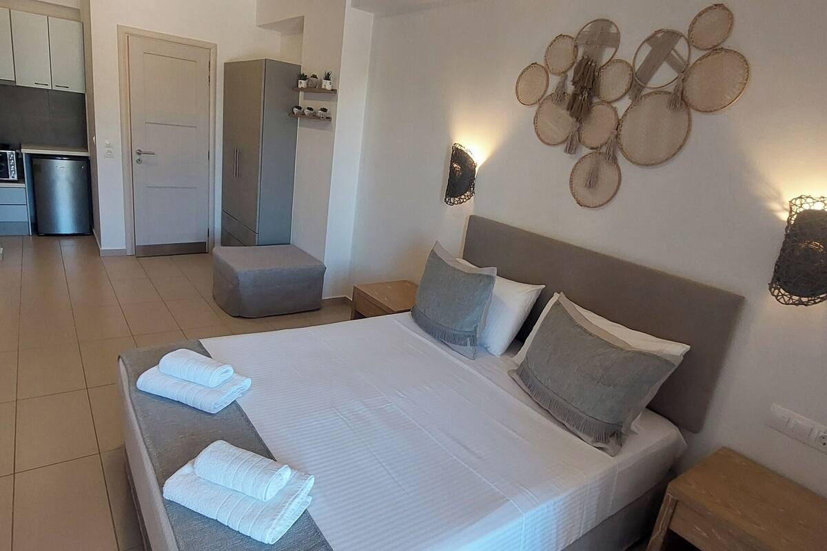 A bedroom with a bed, pillows, and a bedside table. There is a small kitchenette in the background.