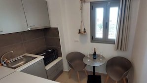 A compact kitchen with a sink, stove, and two chairs around a small table with a bottle and glass. - Selinopetra Rooms (Elafonisos)