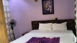 A bedroom with a bed, a wall-mounted picture, a ceiling light, and a floor fan. - Simple And Classic With Affordable Family Bliss At DT-HOMES (ibadan)