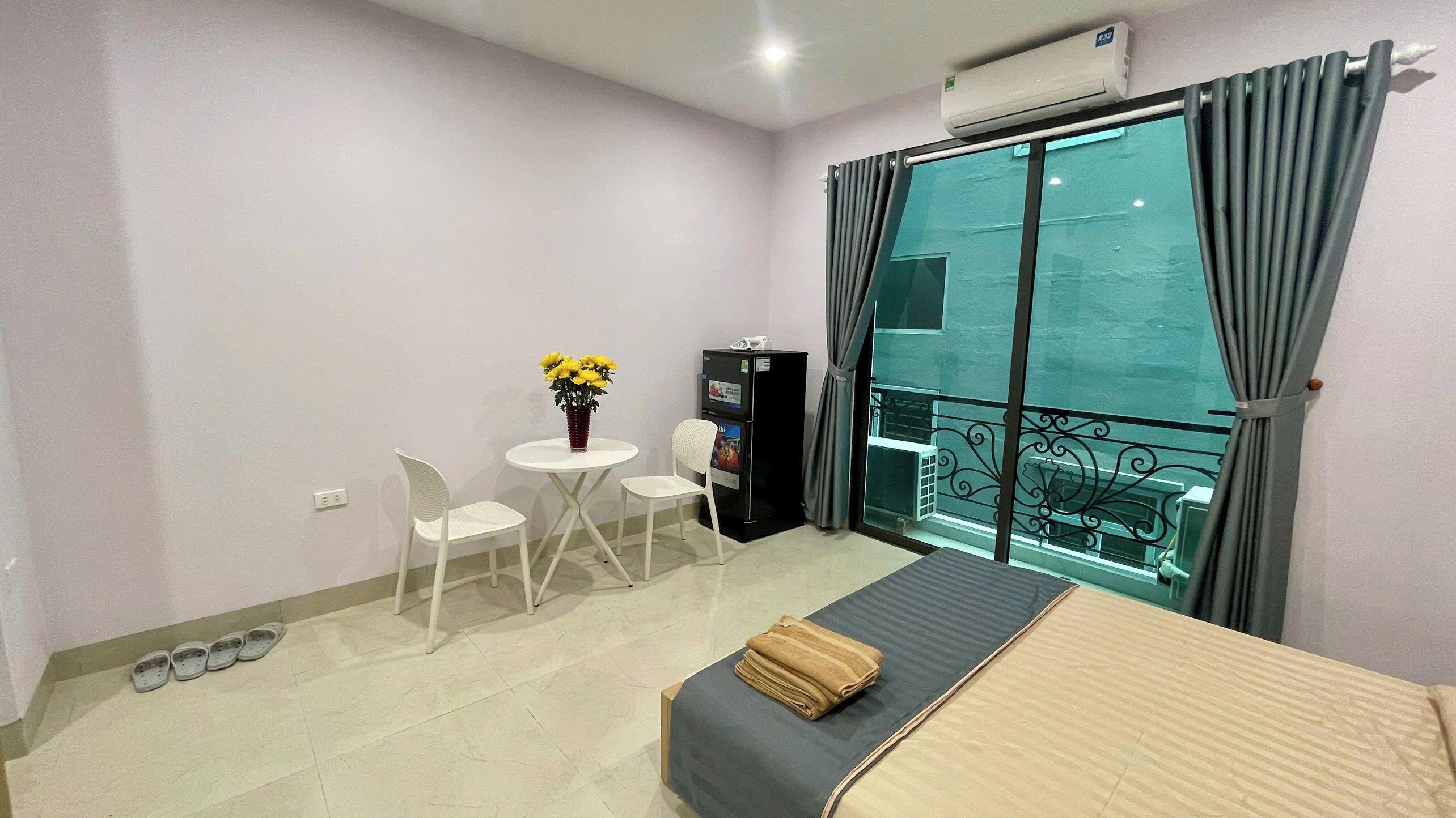 A small, clean room with a bed, a table with chairs, a refrigerator, and a window with curtains.