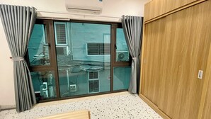Una habitación con un gran ventanal corredero, cortinas grises, un equipo de aire acondicionado, un armario de madera y un piso de terrazo.