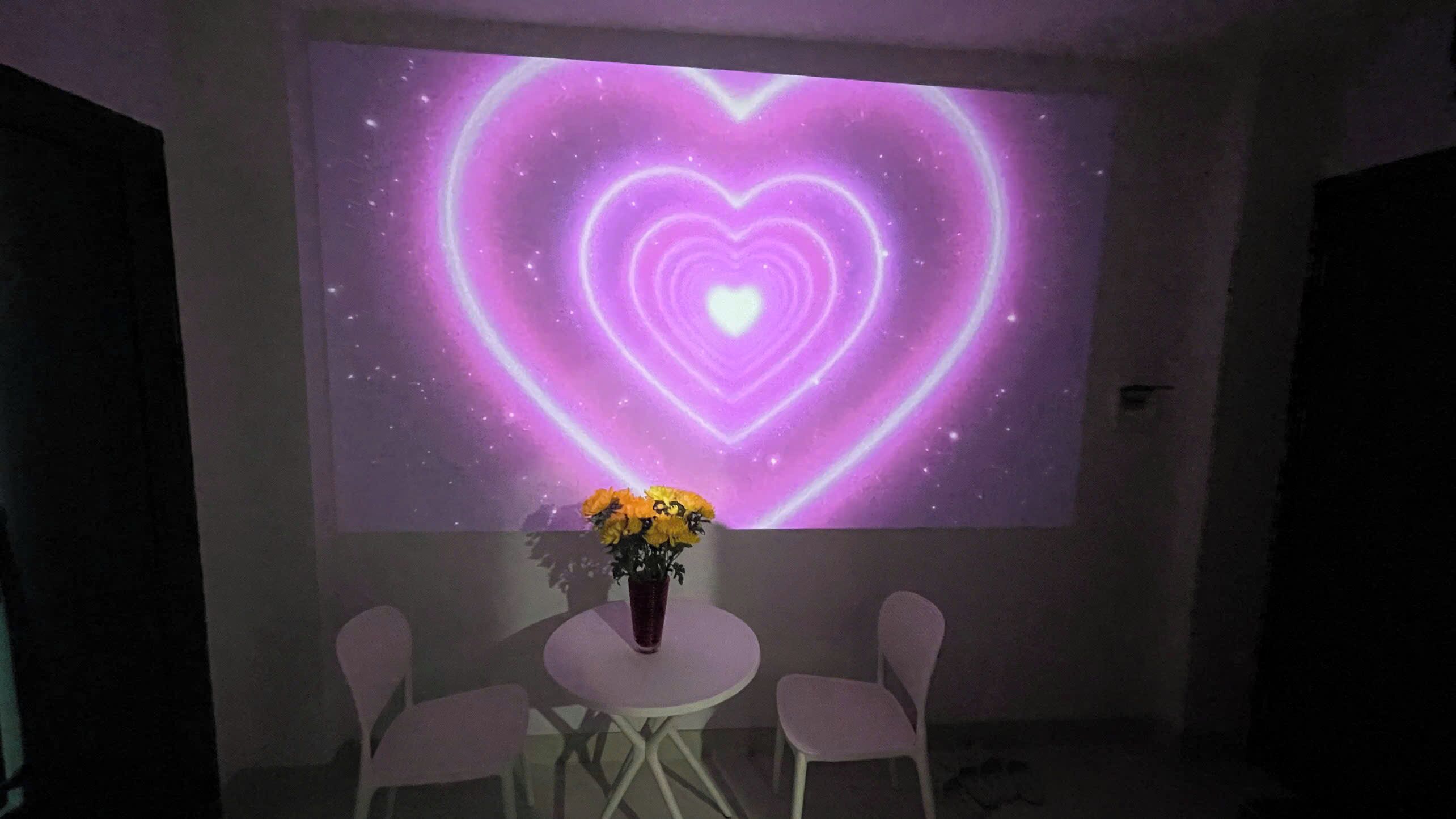 A room with a projection of a heart-shaped design on the wall, a small table with a vase of flowers, and two chairs.